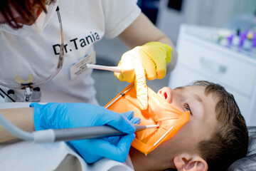 Children's dentistry. First examination at the dentist. A cute handsome boy with his mouth open is looking aside while the doctor is treating her teeth.