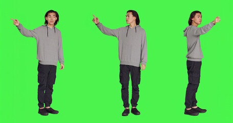 Model showing left or right directions in studio, pointing to both sides and indicating another path. Asian young adult advising someone to go that way, full body greenscreen backdrop.