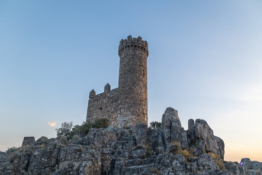 Captures an image of a rock castle with a tower on top, set on rock formations, with a sunset in the background and surrounded by trees.