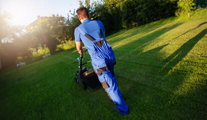 A man in blue work attire mows a green lawn, captured from behind as he works under the warm sunlight. The long shadows and vibrant greenery create a peaceful and productive outdoor scene.