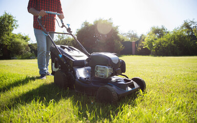Handsome man in a red plaid shirt pushes a lawn mower across a sunlit yard, maintaining the lawn on...