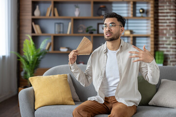 Young man trying to cool down at home using a hand fan, sitting on a couch on a hot day. Relaxed atmosphere despite the heat.