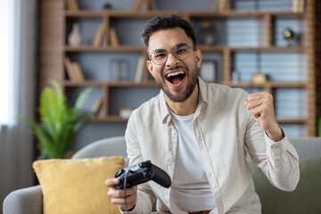 Excited man celebrating video game win at home while holding game controller and smiling on couch