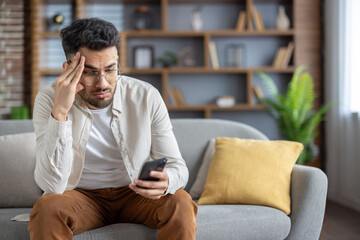 Frustrated man sitting on sofa checking smartphone, feeling stressed at home, wearing casual clothes