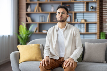 Young man meditating with closed eyes on a comfortable sofa at home, focusing on relaxation and...