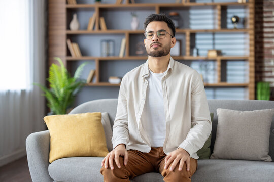 Young man meditating with closed eyes on a comfortable sofa at home, focusing on relaxation and inner peace