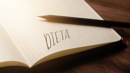 A handwritten inscription "DIETA" on a grille of an open notebook on a wooden countertop, next to a black pencil, lighting of light. (selective focus), translation: Diet
