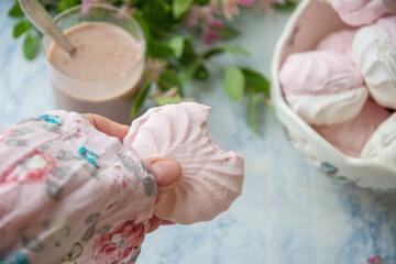 female hand holding bitten vanilla marshmallow,delicate still life with full vase of vanilla marshmallow and glass of coffee with milk, near vase with branches of pink flowers on table,