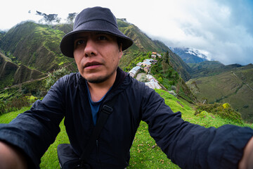 Selfie of a Hispanic traveler in the Andean mountains, wearing a dark hat and black jacket, with a small rural village and green hills under a cloudy sky in the background.