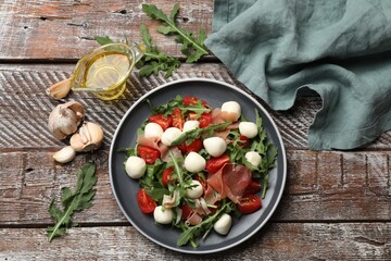 Tasty salad with arugula, mozzarella cheese, tomatoes and jamon on wooden table, flat lay