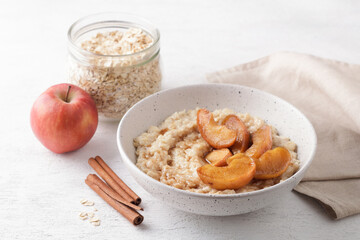 Sweet oatmeal porridge with baked apple slices, cinnamon and caramel sauce in bowl on light background with ingredients