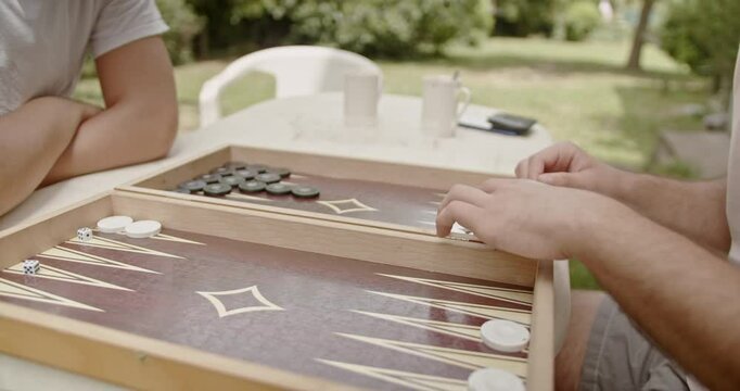 Close-up of two people enjoying a game of backgammon on a sunny day at an outdoor table with coffee mugs.