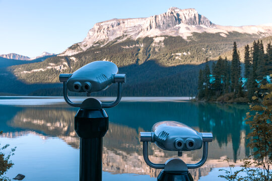 view of a set of public metal binoculars on a stand next to a turquoise lake and a tall mountain in the background on a summer evening