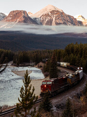 view of a long train with a red locomotive following a curved river on a sunrise with low clouds...