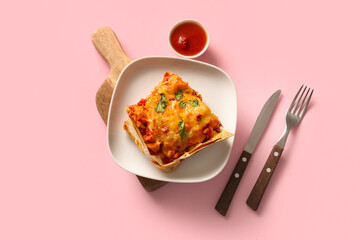 Plate with piece of tasty enchilada, tomato sauce and cutlery on pink background