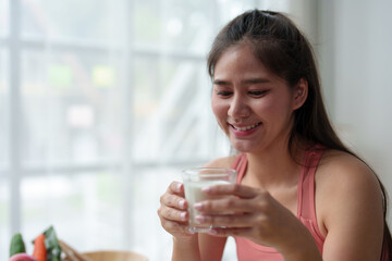 Happy beautiful Asian woman sitting holding a glass of milk after exercise. Fitness at home Healthy woman smiles and drinks protein milk. Vegetable salad. Health and weight loss concept.