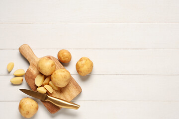 Board with raw potatoes and knife on white wooden background