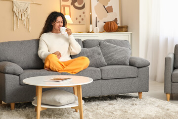 Beautiful young happy African-American woman in autumn outfit with cup of tea sitting on sofa at home