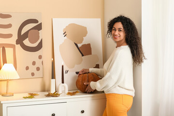 Beautiful young happy African-American woman in stylish outfit with autumn leaves and pumpkin near...