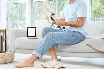 Man with flat feet inserting orthopedic insole into sneaker on sofa at home