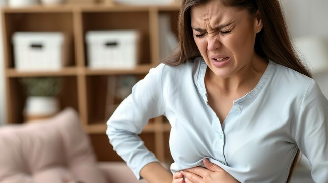 Woman dressed in a light blue shirt clutching below her chest at her home with a pained facial expression. 
