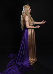 full length portrait of beautiful blonde female model wearing purple and gold silken toga robes with royal crown, historical fantasy goddess costume. Isolated on dark studio background.