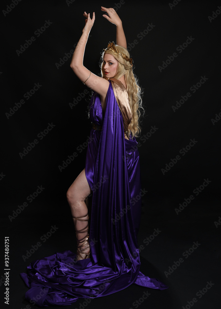 Poster full length portrait of beautiful blonde female model wearing purple and gold silken toga robes with royal crown, historical fantasy goddess costume. Isolated on dark studio background.