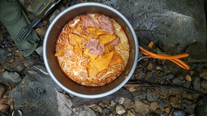 A delicious and unique camping meal featuring a pot of spicy ramen noodles topped with crunchy nachos and sausage slices, cooked outdoors on a rocky surface for an adventurous culinary experience.