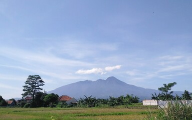 A tranquil view of  salak mountain under a clear blue sky with scattered clouds, surrounded by greenery and rural houses. Ideal for nature, landscape, and peaceful countryside themes.