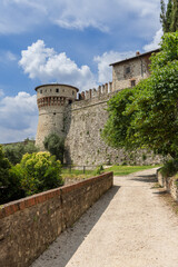 Vertical view of the pathway through Brescia castle park, featuring the famous Torre dei Prigionieri. The image showcases the blend of green foliage and ancient stone architecture