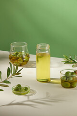On green background with glass flasks full of green olives and olive oil decorated with green leaves. Minimal scene for advertising cosmetic product of olive extract