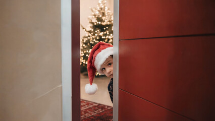 Obraz premium child wearing Santa hat peeking out from the door 