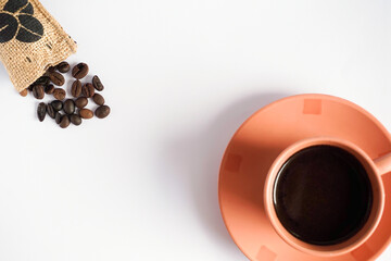 A cup of black coffee placed on the bottom right corner of white background and coffee beans poured out of the bag on top left corner