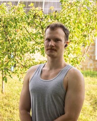 Portrait of a muscular man with mustache and wireless earbuds standing confidently in a green outdoor garden with a solar panel background.