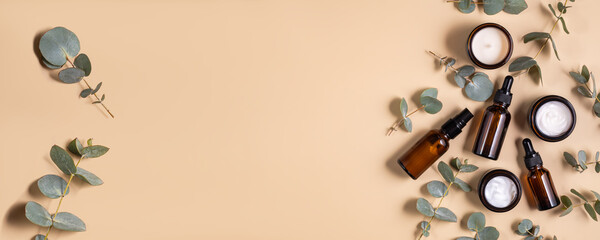 Amber glass essential oil brown bottle and face cream jar with branches of eucalyptus on beige background. Flat lay, top view. Natural organic beauty product packaging mockups. Banner