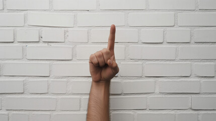 A single hand with the index finger pointing up against a white brick wall background, symbolizing direction or focus