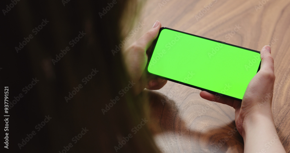 Wall mural young woman sitting at a table and using a smartphone with horizontal green screen