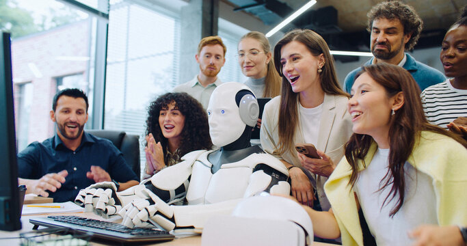 Multi-ethnic group of smart engineers looking at their robot entering text. Checking if program or software operates correctly. Woman next to automated gadget commanding by voice activation.
