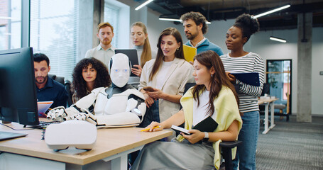 Multi-ethnic group of people surrounding robot. Humanoid working at computer. Colleagues noting everything on tablets and notepads. Woman giving commands. Testing hardware and operating system.
