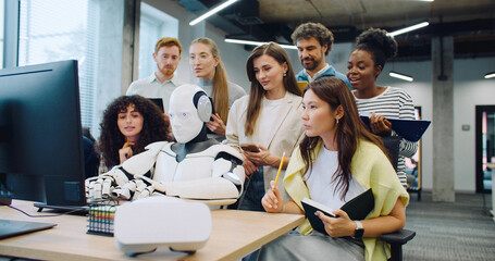 Group of multi-ethnic students surrounding robot. Humanoid sitting at desk and typing on keyboard....