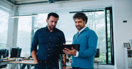Two Caucasian men walking down in middle of long isle of office room. Using tablet device while having discussion. Checking for information online or in database. Management of big business.