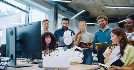 Multi-ethnic colleagues gathering around modern robot. Cyborg sitting at desk with hands on...