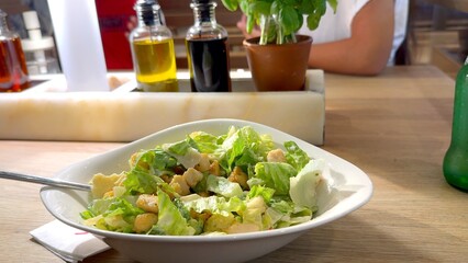 White bowl of Caesar salad sits prominently on table with children playing in lively background of bustling pizzeria Scene blends fresh flavors with fun, family-friendly atmosphere. Basil, olive oil