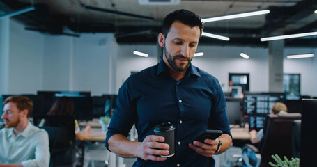 Handsome Caucasian male with dark hair and beard walking down big office space. Holding cup with hot drink with one hand and checking notifications on his phone with other. Walking down between desks.