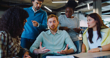 Camera focus on handsome Caucasian man with red hair and moustache. Male trying to explain something to work colleagues. People not agreeing. Difficulty in communication. Negative feedback.