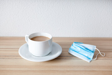 White cup of black coffee with  protective medical mask on wooden  table with empty white background on top