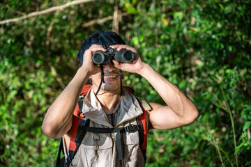 Young Asian man using binoculars exploring forest and enjoying wildlife observation during hiking adventure