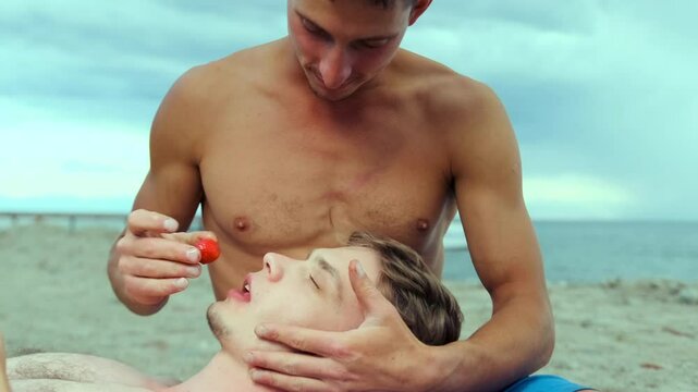 Gay married couple eating strawberries enjoying a sunny day on sandy beach during summer vacation. Slow motion.