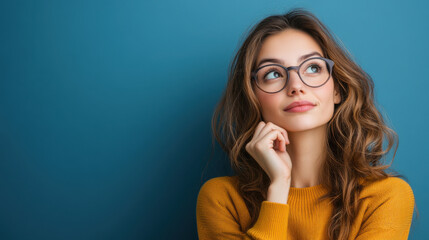 Thoughtful young woman wearing glasses, looking upwards, posing against a blue background, reflecting a contemplative and curious mood.