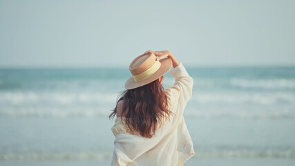 Happy Young Asian woman with straw hat walking on tropical beach, Carefree female enjoying breeze with sea in background. Travel vacation, summer outdoor pleasure.